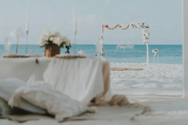Un montaje romántico de propuesta de boda en la playa está capturado en esta imagen. Un arco simple y elegante adornado con flores blancas y tela beige fluyente se encuentra en una playa de arena blanca prístina, con vista a un océano turquesa calmado. Un letrero acrílico claro que dice "Will You Marry Me?" añade un toque de sofisticación moderna. En primer plano, fuera de foco, una mesa bellamente puesta cubierta con lino blanco es visible, completa con centros de mesa florales blancos, sugiriendo una comida celebratoria para seguir la propuesta. La atmósfera general es pacífica, serena y llena de expectativa, enfatizando un sentimiento de amor y felicidad. La iluminación suave y la paleta de colores apagados de blancos, beiges y azules crea una escena soñadora e idílica perfecta para una propuesta memorable.