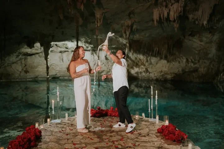 Pareja celebrando con champaña junto al agua turquesa de un cenote de caverna iluminado con velas