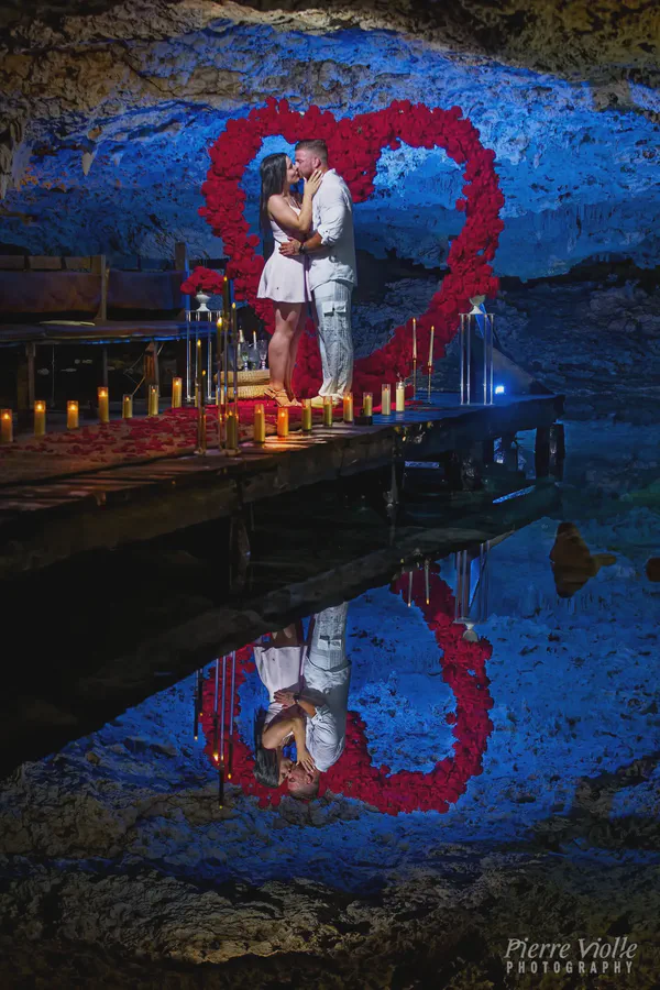 Una pareja romántica comparte un beso apasionado durante una propuesta en un entorno impresionante de cueva. La pareja, vestida de blanco y rosa claro, se encuentra en un pequeño muelle de madera adornado con pétalos de rosa roja y numerosas velas encendidas. Un gran arreglo en forma de corazón de rosas rojas profundas los enmarca contra el telón de fondo de las paredes azules texturizadas de la cueva, creando un ambiente mágico e íntimo. El agua refleja la escena bellamente, reflejando a la pareja y el corazón floral. El ambiente general es de amor, romance y celebración, haciendo de este un momento impresionante e inolvidable capturado en una ubicación única y dramática. La imagen muestra la iluminación dramática, las ricas texturas de la cueva y el contraste vibrante entre las flores rojas y los tonos azules fríos de la cueva.