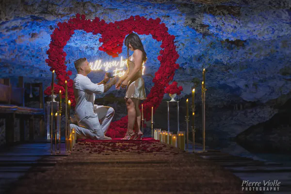 Una escena romántica de propuesta se desarrolla en una cueva débilmente iluminada. Un hombre está arrodillado en una rodilla, presentando un anillo a una mujer que está de pie frente a él. Están posicionados sobre una alfombra roja esparcida con pétalos de rosa, llevando hacia un fondo en forma de corazón adornado con rosas rojas vibrantes. El fondo está iluminado por un letrero de neón que dice "Will you be my forever?". La luz suave de las velas crea un resplandor cálido, reflejándose en los tonos azules fríos de las paredes de la cueva. La mujer lleva un vestido dorado brillante, y el hombre está vestido con una camisa y pantalones blancos nítidos. El ambiente general es íntimo, romántico y lleno de expectativa y alegría. La escena está bellamente montada, capturando un momento mágico e inolvidable.
