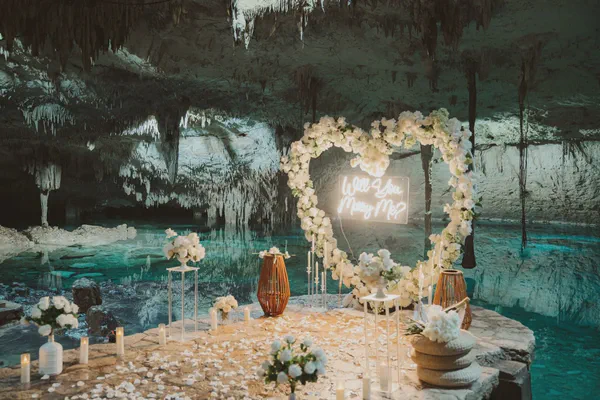 A romantic marriage proposal scene is set inside a stunning cenote cave. A heart-shaped floral arrangement, made of pristine white flowers, frames a glowing neon sign that asks "Will You Marry Me?". The turquoise waters of the cenote reflect the cave's dramatic rock formations, which are adorned with delicate stalactites. Candles and more white floral arrangements are artfully placed around a stone platform, creating a serene and intimate atmosphere. White rose petals scatter the ground, adding to the magical ambiance. The overall color palette is cool and elegant, with the white flowers contrasting beautifully against the teal water and dark cave walls. The scene evokes feelings of wonder, romance, and adventure, making it a truly unforgettable moment.