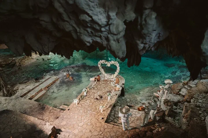 Vista aérea de un arco de corazón y un pasillo con velas junto al agua turquesa de un cenote