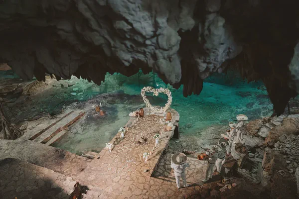 Una vista aérea impresionante de una ceremonia de boda montada dentro de una cueva de cenote deslumbrante. La ceremonia se lleva a cabo en un sendero de piedra que se extiende sobre un cenote turquesa vibrante, con un gran arreglo floral en forma de corazón sirviendo como telón de fondo. Flores blancas y pétalos están esparcidos a lo largo del sendero. Cuatro músicos mariachi en trajes blancos tradicionales y sombreros de ala ancha se encuentran en los escalones de piedra, añadiendo al ambiente mágico. El techo oscuro y rocoso de la cueva proporciona un contraste dramático con el agua brillante y clara y la decoración floral blanca suave. La escena general es romántica, mística y evocadora de una celebración de boda única e inolvidable en un hermoso entorno natural. La iluminación acentúa la interacción dramática de luz y sombra, realzando el ambiente mágico y algo misterioso.