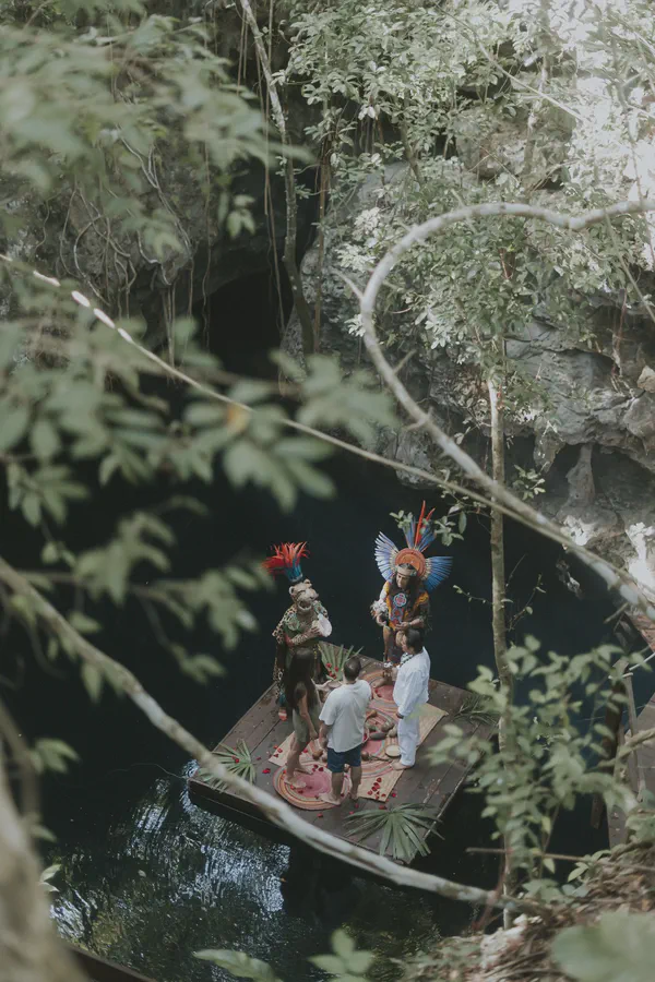Una vista aérea de una ceremonia de boda maya que se lleva a cabo en una pequeña plataforma de madera sobre un cenote oscuro y quieto (sumidero). Dos individuos en ropa maya tradicional vibrante, adornados con elaborados tocados de plumas rojas, azules y doradas, presiden la ceremonia. Una pareja se encuentra descalza ante ellos, vestida con ropa simple blanca y verde, participando en el ritual. La plataforma está decorada con una alfombra circular en tonos tierra y pétalos de rosa roja esparcidos. El follaje tropical verde exuberante rodea el cenote, enmarcando la escena y añadiendo a la atmósfera mística. El ambiente general es de reverencia y espiritualidad, los verdes profundos y azules contrastando bellamente con los colores brillantes del atuendo maya. La imagen captura un evento único y culturalmente significativo ambientado contra el telón de fondo impresionante de una maravilla natural.