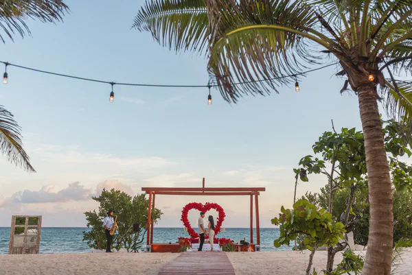 Una romántica propuesta en la playa se desarrolla al atardecer en el Caribe. Una pareja se encuentra de la mano ante un gran arreglo floral en forma de corazón hecho de rosas rojas bajo una pérgola de madera. La escena está iluminada por cálidas luces de cadena colgadas entre palmeras. Un saxofonista es visible a la izquierda, añadiendo a la atmósfera íntima. El océano turquesa calmado proporciona un telón de fondo impresionante, y los suaves colores pastel del atardecer se reflejan en la arena. El ambiente general es de amor, alegría y celebración. La pareja parece feliz y relajada mientras esperan la ocasión trascendental. El entorno se siente lujoso y aislado.