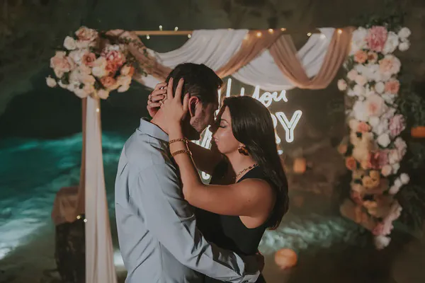 Una propuesta romántica de compromiso se desarrolla en un impresionante entorno de cueva. Un hombre y una mujer, profundamente enamorados, se abrazan y comparten un momento tierno, con sus frentes tocándose. La mujer, con cabello largo y oscuro y un vestido negro, tiene sus brazos alrededor del cuello del hombre. Él lleva una camisa azul claro. Detrás de ellos, un hermoso arco floral decorado con flores de colores pastel y iluminación suave crea un ambiente mágico. Un letrero de neón que dice "Will You Marry Me?" añade a la ocasión especial. Las paredes de la cueva son oscuras, contrastando con el agua de color turquesa visible en el fondo, realzando la atmósfera romántica e íntima de la escena. El sentimiento general es de alegría, amor y emoción mientras la pareja celebra esta ocasión trascendental.