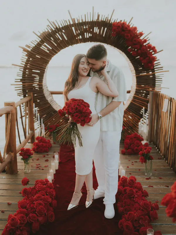 Una imagen conmovedora captura una escena romántica de propuesta. Una pareja, vestida con elegante ropa blanca, se encuentra abrazada bajo un arco circular rústico de madera adornado con rosas rojas vibrantes. La mujer sostiene un gran ramo de rosas rojas profundas, su sonrisa radiante mientras su pareja se acerca. El entorno es una hermosa terraza al aire libre con vista a un cuerpo de agua tranquilo, creando una atmósfera serena e íntima. Una alfombra roja lujosa lleva a la pareja, flanqueada por más rosas rojas en cascada y luz suave de velas. La paleta de colores general es rica y romántica, enfatizando la conexión apasionada entre la pareja y la ocasión alegre. La imagen irradia amor, felicidad y la emoción de un momento especial. Los detalles, desde los impresionantes arreglos florales hasta las expresiones felices de la pareja, bellamente cuentan la historia de una propuesta memorable.