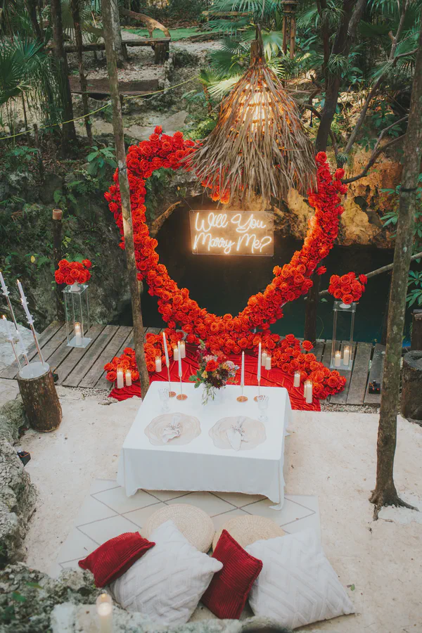 Una escena romántica de propuesta está ambientada en una selva exuberante. Un arreglo en forma de corazón de rosas rojas vibrantes forma un telón de fondo para un letrero de neón que dice "Will You Marry Me?". Una mesa pequeña e íntima está puesta para dos, cubierta con un mantel blanco y adornada con velas y elegantes servicios de mesa. Cojines suaves de color crema y esteras tejidas proporcionan asientos cómodos en el suelo arenoso. La atmósfera general es cálida, íntima y llena de amor y expectativa. La escena está iluminada por el resplandor suave de las velas y el letrero de neón, contrastando bellamente contra los verdes profundos y marrones del follaje tropical. El entorno sugiere un momento de propuesta aislado y mágico en un paraíso tropical.