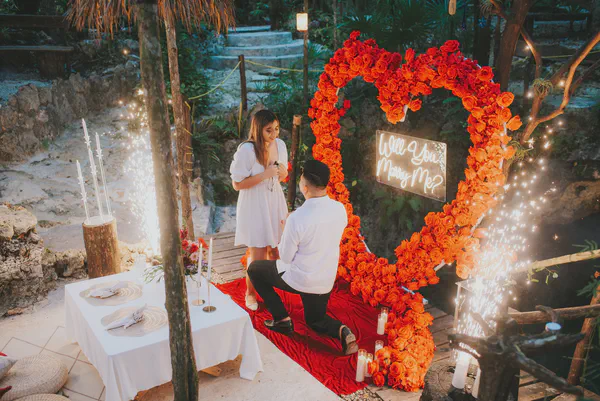 Una escena romántica de propuesta está ambientada en una exuberante selva tropical. Un impresionante arreglo floral en forma de corazón, compuesto completamente de rosas rojas vibrantes, sirve como un telón de fondo dramático. El corazón está bellamente iluminado y enmarcado por la densa vegetación verde, creando un contraste sorprendente entre las flores rojas intensas y el follaje natural. La escena está cuidadosamente compuesta para evocar romance y pasión, con la naturaleza proporcionando un ambiente íntimo y privado. La iluminación suave realza los colores ricos y crea una atmósfera mágica perfecta para un momento de propuesta inolvidable en un paraíso tropical escondido.