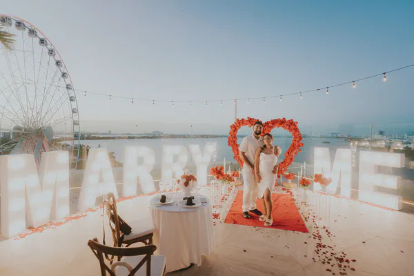 Una romántica propuesta en la azotea se desarrolla bajo un cielo crepuscular. Una pareja, vestida con elegante ropa blanca, se encuentra orgullosamente ante letras iluminadas "Marry Me". Posan en medio de un arreglo floral en forma de corazón de rosas coral vibrantes, creando un telón de fondo contra una vista panorámica de la ciudad y una noria. Pétalos de rosa roja esparcidos adornan el suelo, llevando a una mesa bellamente puesta para dos. La iluminación suave de luces de cadena y velas añade al ambiente íntimo, capturando un momento de alegría y amor. El ambiente general es cálido, romántico y celebratorio.