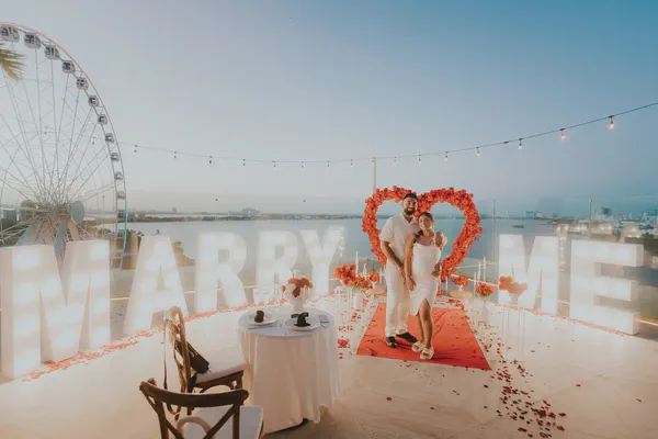 A romantic rooftop proposal unfolds under a twilight sky. A couple, dressed in elegant white attire, stands proudly before illuminated "Marry Me" letters. They pose amidst a heart-shaped floral arrangement of vibrant coral roses, creating a backdrop against a panoramic city and Ferris wheel view. Scattered red rose petals adorn the floor, leading to a beautifully set table for two. Soft lighting from string lights and candles adds to the intimate ambiance, capturing a moment of joy and love. The overall mood is warm, romantic, and celebratory.