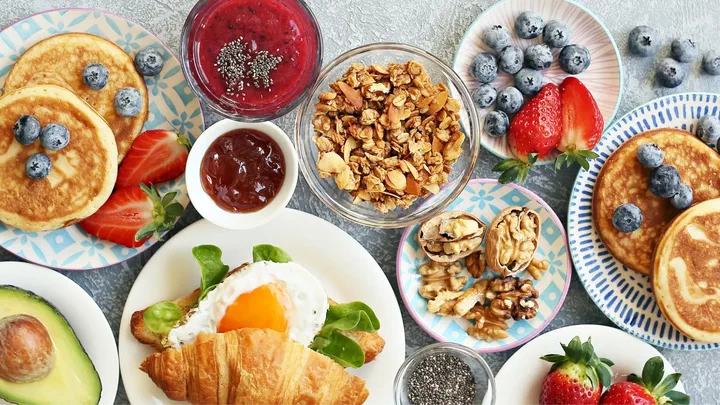 Pancakes with berries, a croissant with a fried egg, avocado, nuts and jam spread across a brunch table.