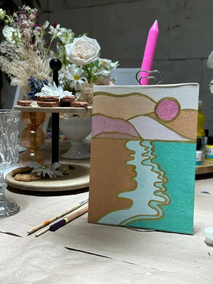 A finished canvas of a golden beach and turquoise sea stands on a table beside flowers, a candle and a tray of cookies.