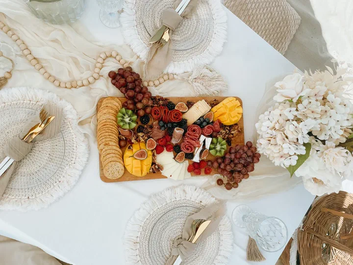 Vista desde arriba: una tabla de charcutería con fruta, quesos, carnes frías y galletas entre manteles individuales con flecos, cubiertos dorados y flores blancas.